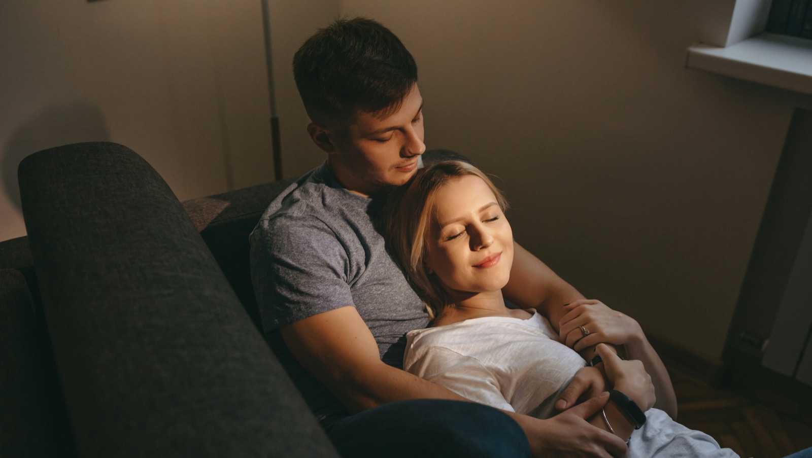 Pareja en sofá con manos sobre vientre de embarazada · luz cálida íntima